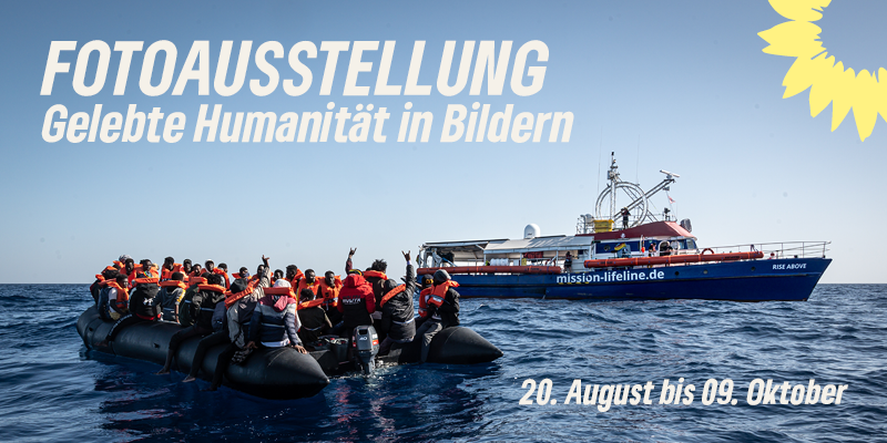 Eine Rettungsmission von Mission Lifeline auf dem offenen Meer. Im Hintergrund ein Boot des Vereins, im Vordergrund ein Schlauchboot voller Menschen mit Rettungswesten. Folgender Text ist auf dem Bild: "Fotoausstellung" "Gelebte Humanität in Bildern" "20. August bis 09. Oktober" Zudem in der Ecke oben rechts die BÜNDNISGRÜNE Sonnenblume