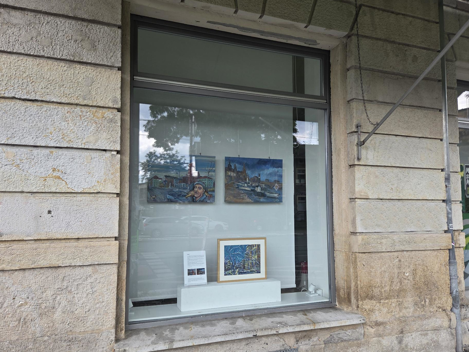Im Schaufenster hängen zwei gemallte Bilder. Eines zeigt den Königsbrücker Platz im Hechtviertel. Das andere eine Ansicht vom Elbufer auf die Hofkirche und das Schloß. Auf einem Sockel steht ein weiteres Bild, welches eine alte Villa in der Neustadt zeigt.