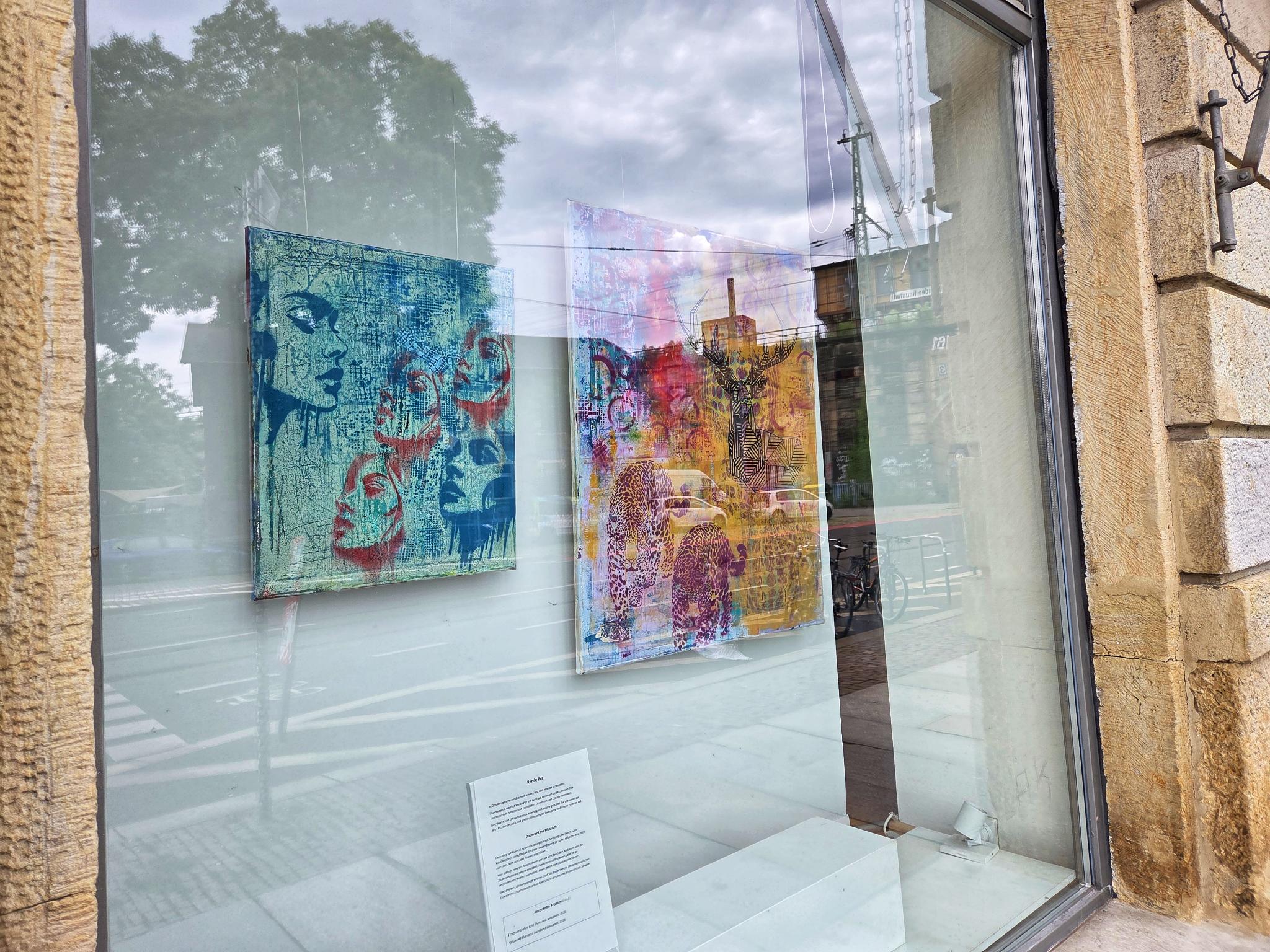 Zwei Bilder hängen im Kunstfenster. Sie sind farbenfroh gestaltet. Das eine zeigt verschiedene weibliche Köpfe vor hellblauem Hintergrund. Auf dem zweiten sind ein Hirsch und zwei Raubkatzen zu erkennen.